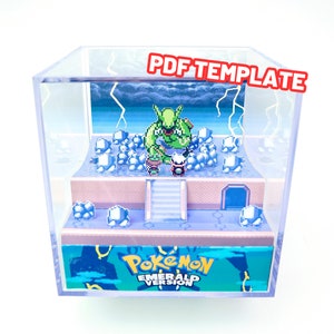 Może przedstawiać: Przezroczysta akrylowa skrzynka z trójwymiarową dioramą papierową w środku. Diorama przedstawia scenę z gry wideo Pokemon Emerald, z zielonym, pikselowanym Pokemonem otoczonym lodem i błyskawicami. Podstawa dioramy ma nadrukowany tekst "Pokemon Emerald Version".