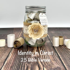 May include: A glass jar with a silver lid, filled with rolled-up papers. The jar is decorated with burlap, lace, and a paper flower. The text "Identity in Christ 25 Bible Verses" is displayed on the jar. A tag reads "Be encouraged and reminded by these 25 biblical truths of who you really are."