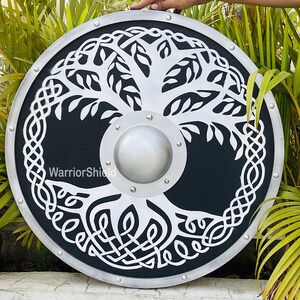 May include: A circular shield in black and silver, featuring a white tree of life motif. The shield has a silver edge and a central boss. The text "WarriorShield" is visible. The shield is displayed against a backdrop of green foliage.