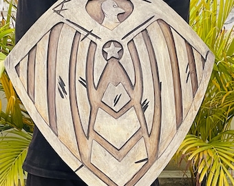 May include: A wooden shield with a carved design featuring an eagle, star, and geometric shapes. The shield has a light brown colour with darker brown accents, and is held in front of a person.