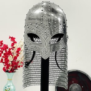 May include: A silver Viking helmet with intricate etched designs and a chainmail aventail. The helmet is on a black stand and has the text "WarriorShield" on the front.