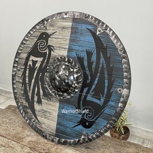 May include: A round wooden shield with a black metal rim and a black and white design of two stylized birds. The shield is painted with a blue and white background and has the text "WarriorShield" printed on it.