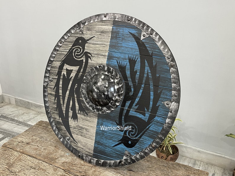 May include: A round wooden shield with a black metal rim and a black and white design of two stylized birds. The shield is painted with a blue and white background and has the text "WarriorShield" printed on it.