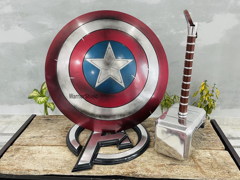 May include: A Captain America shield with a star in a blue circle, surrounded by red and silver rings. Next to it is Thor's hammer with a brown handle and a silver head. The shield is on a stand with the Avengers logo.