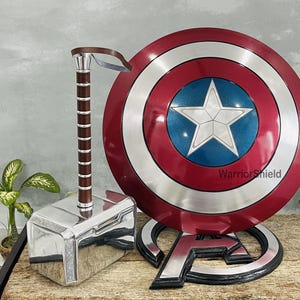 May include: A silver and brown hammer, a red, white, and blue shield with a white star, and a silver Avengers logo. The shield has the text "WarriorShield". The items are displayed on a wooden surface.