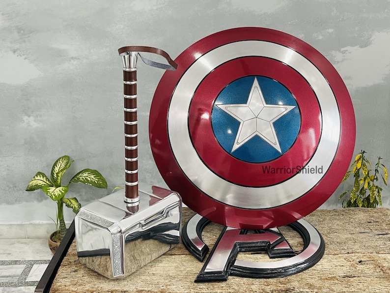 May include: A silver and brown hammer, a red, white, and blue shield with a white star, and a silver Avengers logo. The shield has the text "WarriorShield". The items are displayed on a wooden surface.