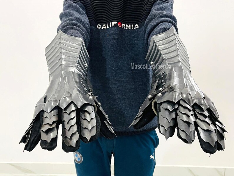 May include: A pair of black metal gauntlets with a scaly design. The gauntlets are worn by a person wearing a blue sweater with the word "CALIFORNIA" on it.