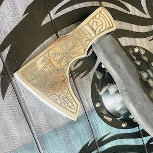 May include: A silver axe with a wooden handle and intricate etched designs. The axe head has a Celtic knot design and the blade is decorated with a floral pattern. The axe is resting on a wooden shield with a black and silver design.