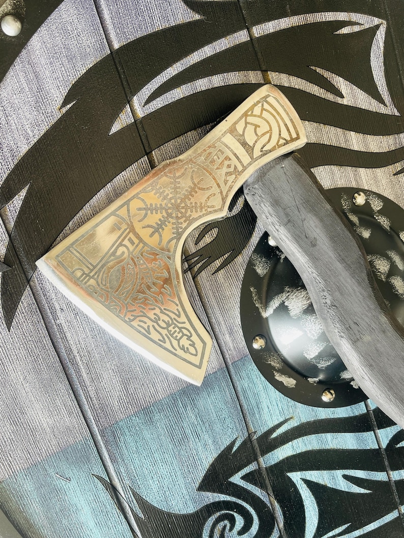 May include: A silver axe with a wooden handle and intricate etched designs. The axe head has a Celtic knot design and the blade is decorated with a floral pattern. The axe is resting on a wooden shield with a black and silver design.