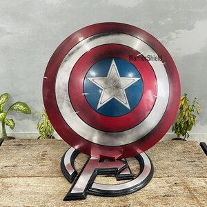May include: A Captain America shield replica, featuring a red, white, and blue color scheme with a central white star on a blue field. The shield is displayed on a black and silver stand with the Avengers logo. The text "WarriorShield" is visible.