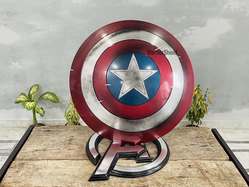 May include: A Captain America shield replica, featuring a red, white, and blue color scheme with a central white star on a blue field. The shield is displayed on a black and silver stand with the Avengers logo. The text "WarriorShield" is visible.