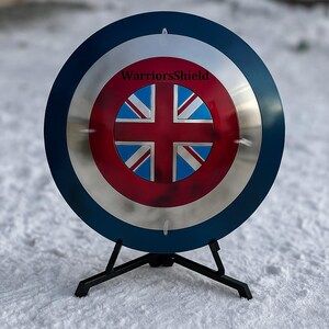 May include: A round shield with a blue border, silver center, and a red and blue Union Jack design. The text "WarriorsShield" is printed on the top. The shield is mounted on a black stand.