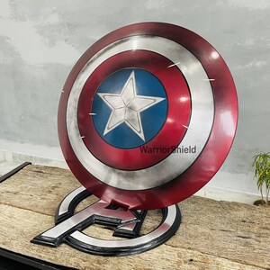 May include: A Captain America shield replica, featuring a red, white, and blue color scheme with a central white star on a blue background. The shield is displayed on a black and silver Avengers logo stand. The text "Warrior Shield" is visible on the shield.