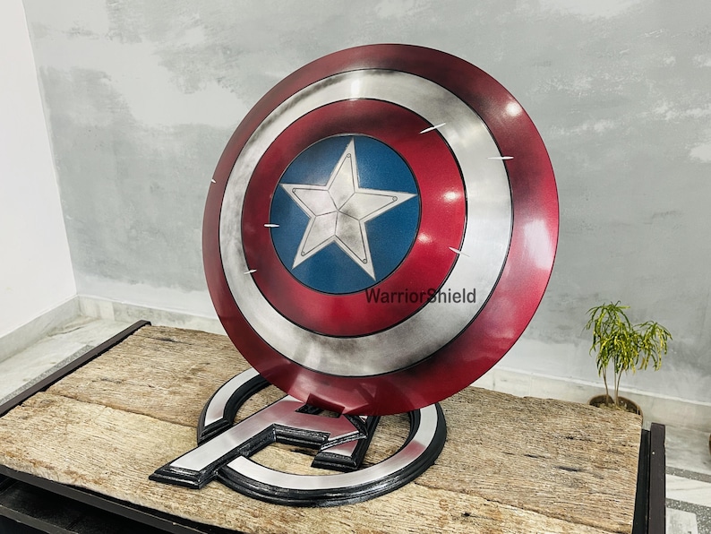May include: A Captain America shield replica, featuring a red, white, and blue color scheme with a central white star on a blue background. The shield is displayed on a black and silver Avengers logo stand. The text "Warrior Shield" is visible on the shield.