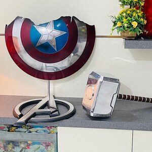 May include: A silver and black replica of Thor's hammer, Mjolnir, with a brown leather strap. A silver Captain America shield with a red, white, and blue design is on a black stand with the Avengers logo.