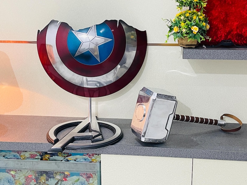 May include: A silver and black replica of Thor's hammer, Mjolnir, with a brown leather strap. A silver Captain America shield with a red, white, and blue design is on a black stand with the Avengers logo.
