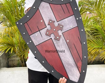 May include: A red and white wooden shield with a black leather trim and metal rivets. The shield has a cross design with a metal center and the text "WarriorShield" printed on the front.