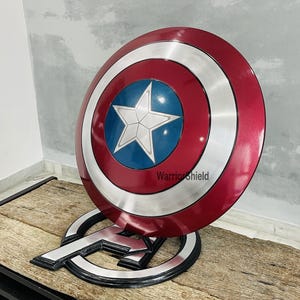 May include: A Captain America shield replica, featuring a red, white, and blue color scheme. The shield has a central blue circle with a white star, surrounded by a silver ring and a red outer ring. The shield is displayed on a stand with the Avengers logo.