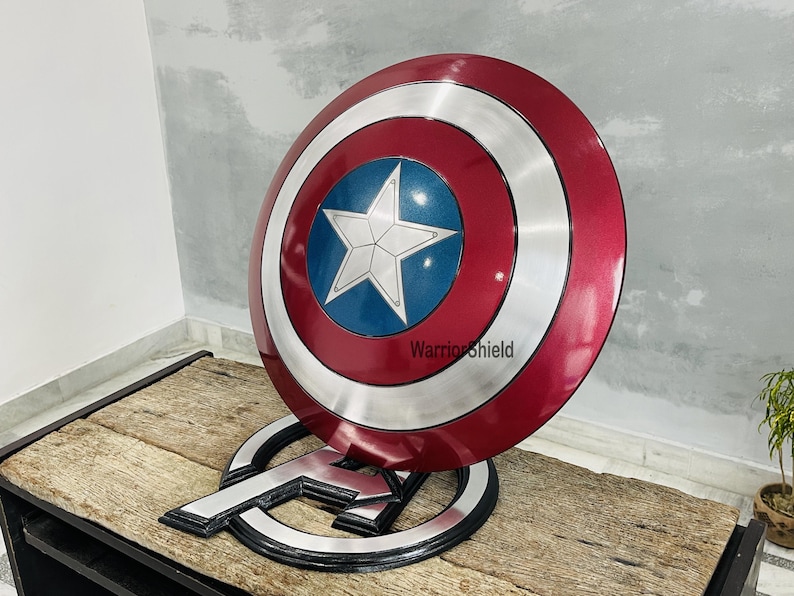 May include: A Captain America shield replica, featuring a red, white, and blue color scheme. The shield has a central blue circle with a white star, surrounded by a silver ring and a red outer ring. The shield is displayed on a stand with the Avengers logo.