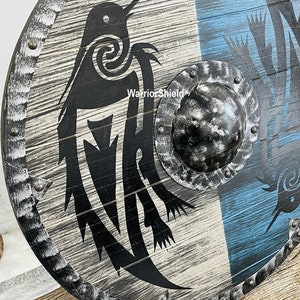 May include: A round wooden shield with a black and white design of two birds. The shield has a metal rim with rivets and a metal boss in the center. The shield is painted with a blue and white background.