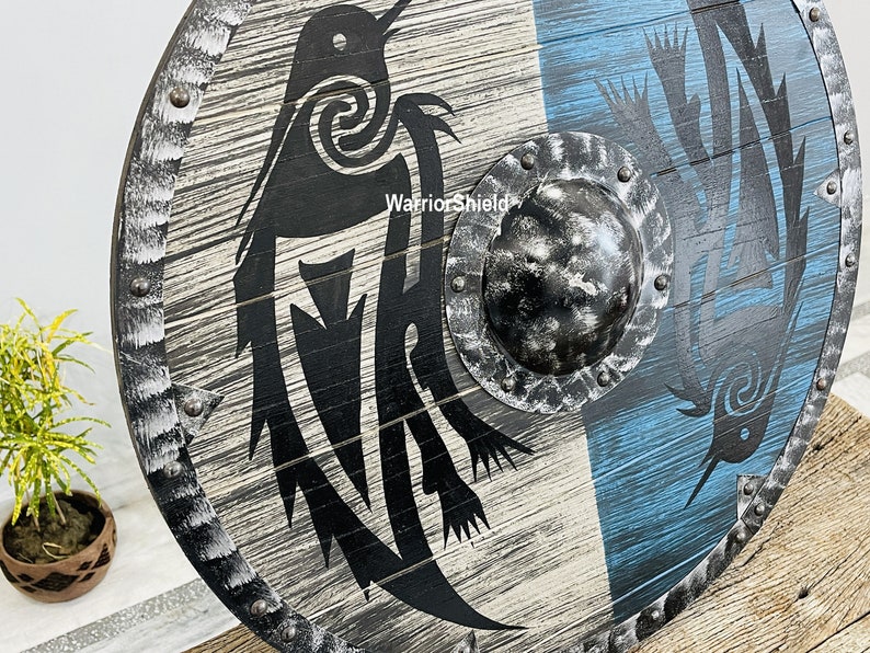 May include: A round wooden shield with a black and white design of two birds. The shield has a metal rim with rivets and a metal boss in the center. The shield is painted with a blue and white background.