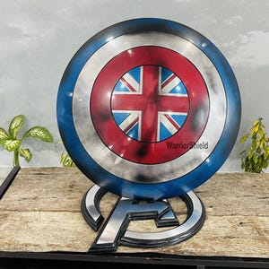 May include: A blue, red, and white Captain America shield with a Union Jack design in the center. The shield is on a stand with the letter "A" on it. The text "WarriorShield" is visible on the shield.