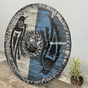 May include: A round wooden shield with a black and white design of two birds. The shield has a blue and white background and a metal rim with rivets. The text "WarriorShield" is printed on the shield.