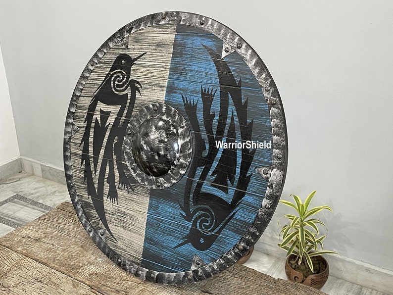 May include: A round wooden shield with a black and white design of two birds. The shield has a blue and white background and a metal rim with rivets. The text "WarriorShield" is printed on the shield.