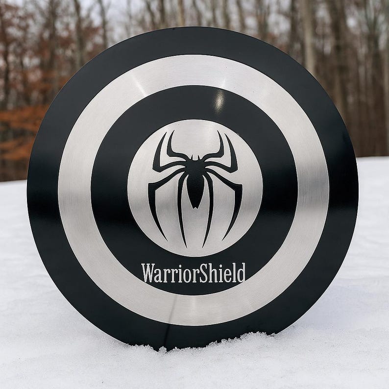 Puede incluir: Un escudo negro y plateado con un emblema de ara&ntilde;a en el centro. El escudo tiene c&iacute;rculos conc&eacute;ntricos y el texto "WarriorShield" en la parte inferior. El escudo es redondo y parece ser de metal.