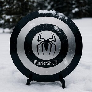 Puede incluir: Un escudo negro y plateado con un emblema de ara&ntilde;a y el texto "WarriorShield". El escudo circular, con anillos conc&eacute;ntricos, est&aacute; sobre un soporte negro en la nieve.