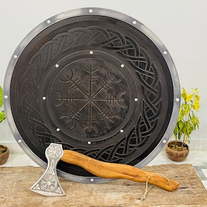 May include: A black wooden shield with a silver trim and a carved Viking compass design. A silver axe with a wooden handle and a Viking compass design is lying on a wooden surface in front of the shield.