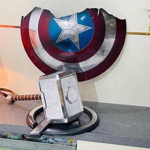 May include: A silver and gold replica of Mjolnir, Thor's hammer, with a brown leather strap handle, resting on a black circular stand. The hammer is in front of a red, white and blue Captain America shield.