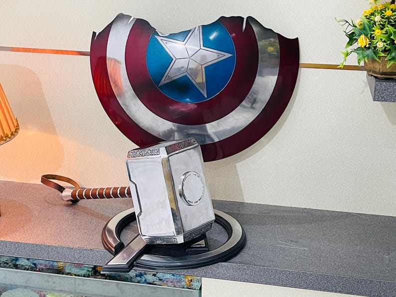 May include: A silver and gold replica of Mjolnir, Thor's hammer, with a brown leather strap handle, resting on a black circular stand. The hammer is in front of a red, white and blue Captain America shield.
