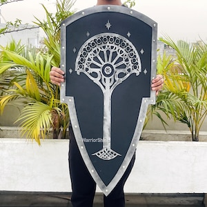 May include: A black and silver shield featuring a stylised tree design and stars. The shield is being held by a person in front of a green leafy background.