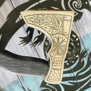 May include: A silver axe with a black wooden handle. The axe head is decorated with intricate engravings of Norse symbols and designs.