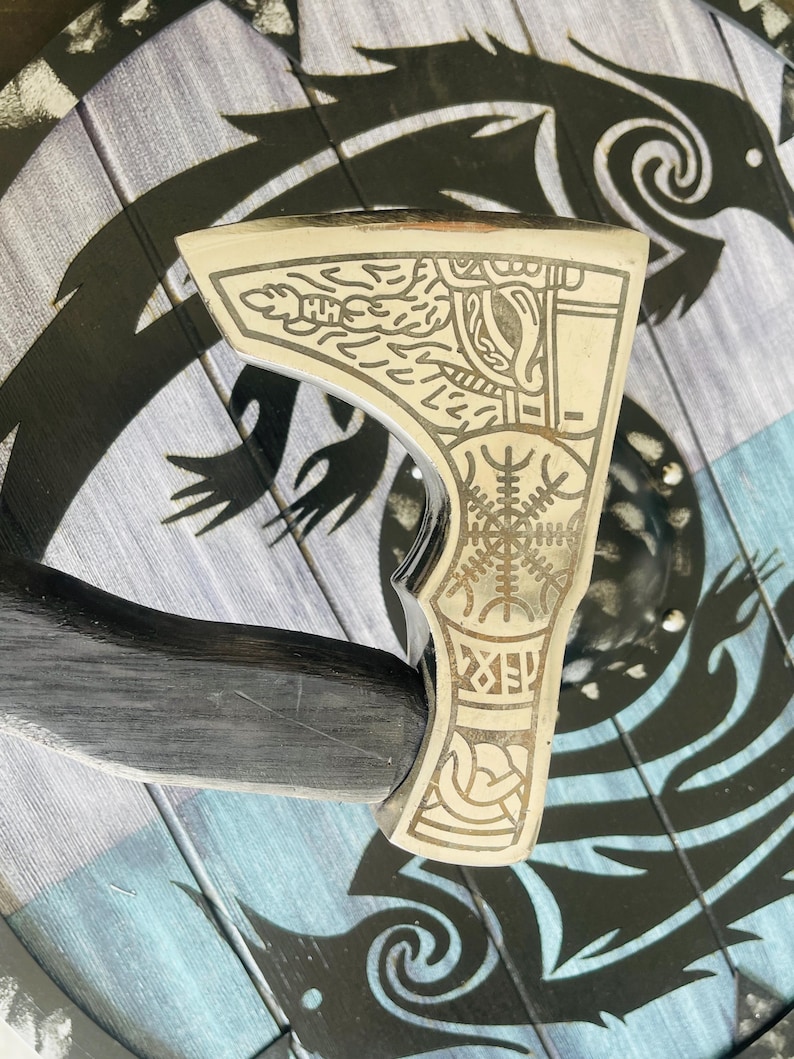 May include: A silver axe with a black wooden handle. The axe head is decorated with intricate engravings of Norse symbols and designs.