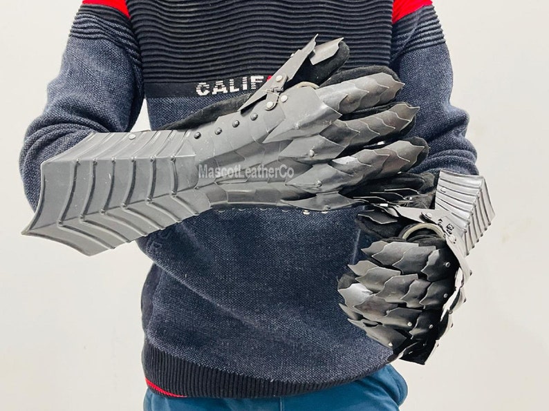 May include: A pair of black metal gauntlets with a claw-like design. The gauntlets are worn by a person wearing a black and red sweater. The text "Mascott LeatherCo" is visible on the gauntlets.