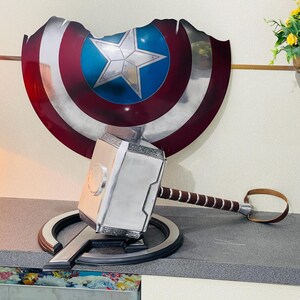 May include: A silver and black replica of Thor's hammer, Mjolnir, with a brown leather strap. The hammer is resting on a silver and black replica of Captain America's shield.