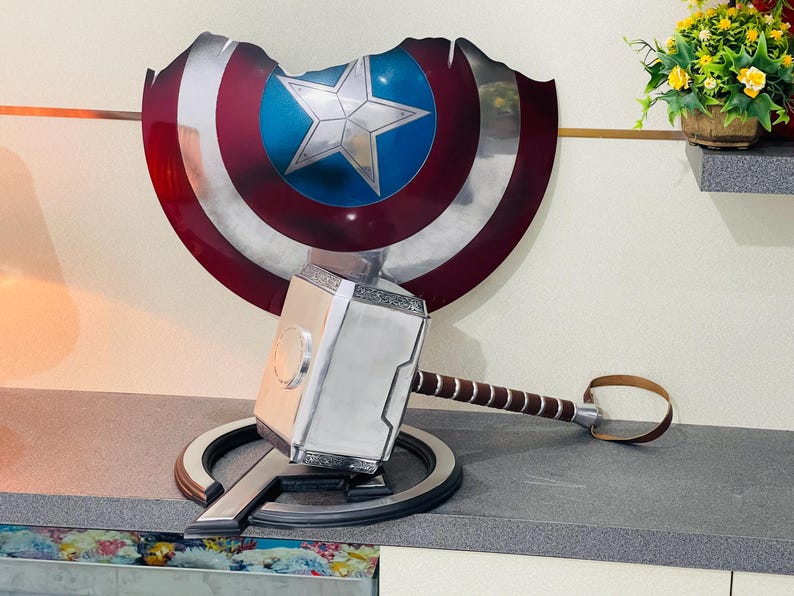 May include: A silver and black replica of Thor's hammer, Mjolnir, with a brown leather strap. The hammer is resting on a silver and black replica of Captain America's shield.