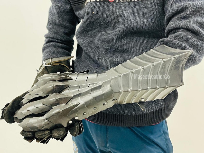 May include: A pair of black metal gauntlets with a dragon scale design. The gauntlets are worn on a person's hands and forearms. The gauntlets are made of metal and have a textured surface. The gauntlets are designed to protect the wearer's hands and forearms from injury. The gauntlets are a popular choice for LARP and cosplay. The gauntlets are also a popular choice for collectors of medieval armor. The gauntlets are made by MascotleatherCo.