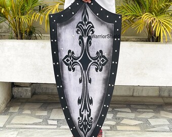 May include: A black and silver shield with a decorative cross design. The shield is made of metal and has a leather strap. The shield is held by a person's hands. The text "WarriorShield" is visible on the shield.