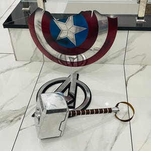 May include: A silver Thor's hammer with a brown leather handle, and a red, white and blue Captain America shield on a stand.