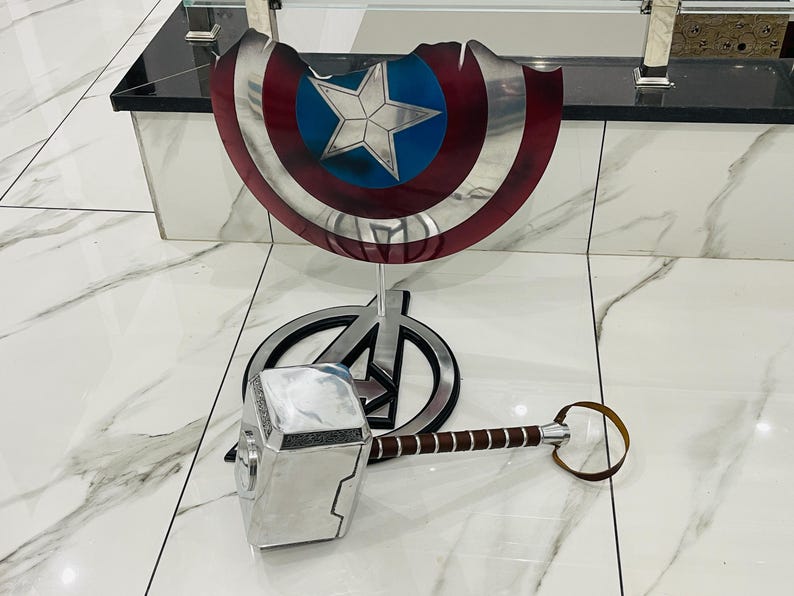May include: A silver Thor's hammer with a brown leather handle, and a red, white and blue Captain America shield on a stand.