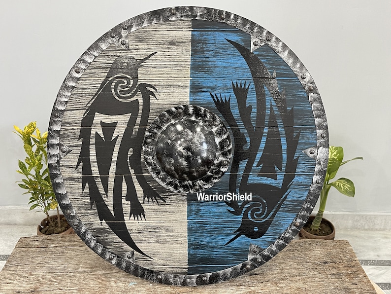 May include: A round wooden shield with a black and white design on one side and a blue and black design on the other side. The shield has a metal rim with rivets and a metal center boss. The text "WarriorShield" is printed on the shield.