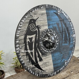 May include: A round wooden shield with a black and white design of a bird on one side and a blue and white design on the other side. The shield has a metal rim with rivets and a metal center boss. The shield is labeled "WarriorShield".
