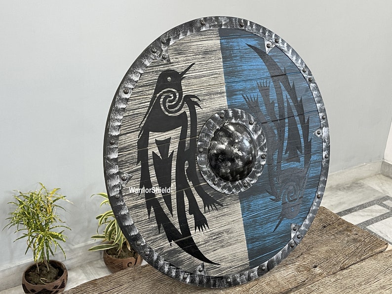 May include: A round wooden shield with a black and white design of a bird on one side and a blue and white design on the other side. The shield has a metal rim with rivets and a metal center boss. The shield is labeled "WarriorShield".