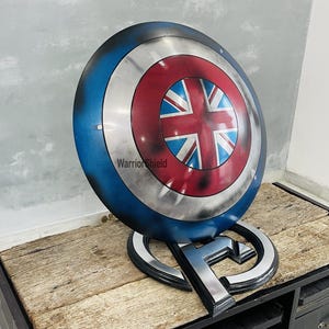 May include: A blue, red, and white Captain America shield with a Union Jack design in the center. The shield is on a stand with the letter "A" on it. The text "Warrior Shield" is visible on the shield.