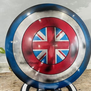May include: A round, blue and silver Captain America shield with a red, white, and blue Union Jack design in the center. The shield has a distressed, weathered look. The text "WarriorShield" is printed on the shield.