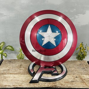 May include: A Captain America shield replica, featuring a red, white, and blue color scheme. The shield has a central blue circle with a white star, surrounded by concentric silver and red rings. It rests on an Avengers logo stand.