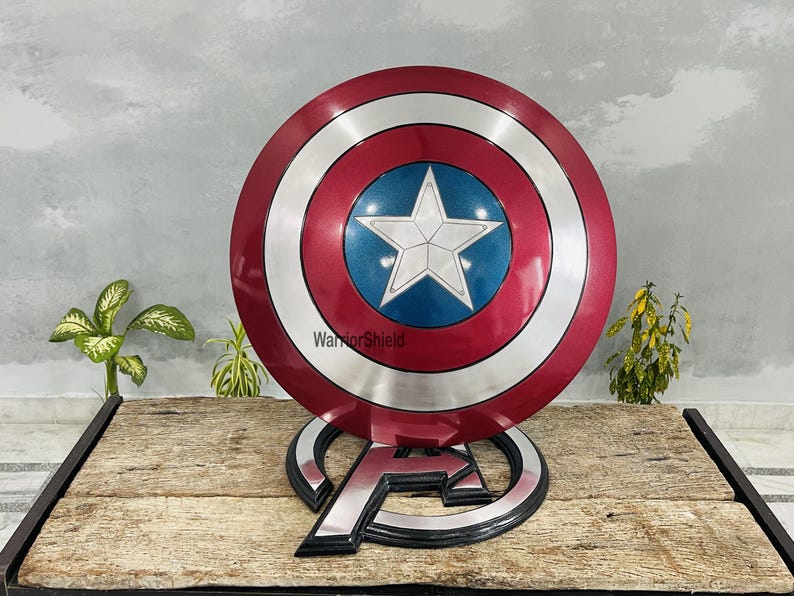 May include: A Captain America shield replica, featuring a red, white, and blue color scheme. The shield has a central blue circle with a white star, surrounded by concentric silver and red rings. It rests on an Avengers logo stand.